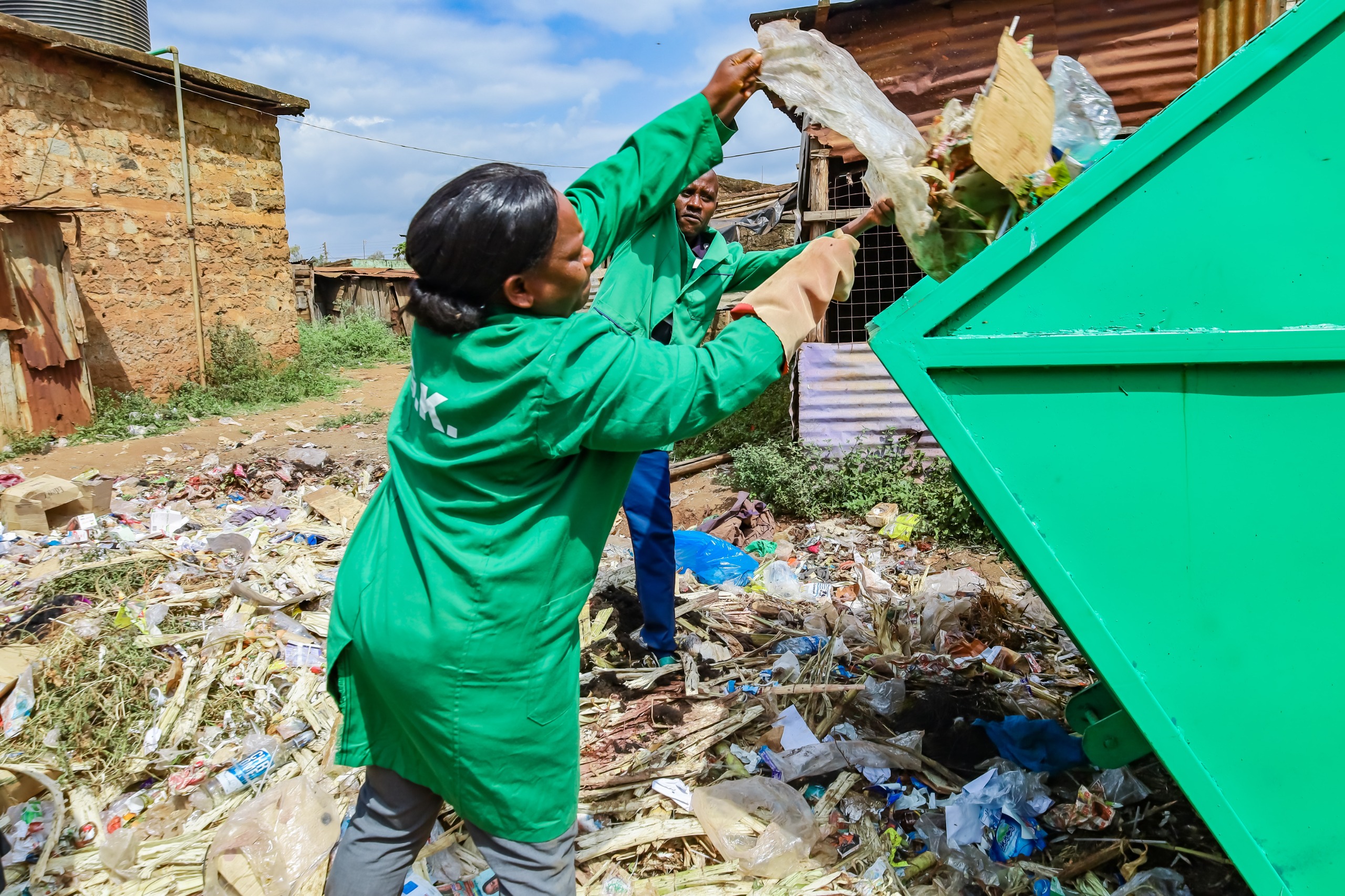 wo workers throwing trash into a metal skip provided by the Kirinyaga County Government. They are cleaning up an area with scattered waste.