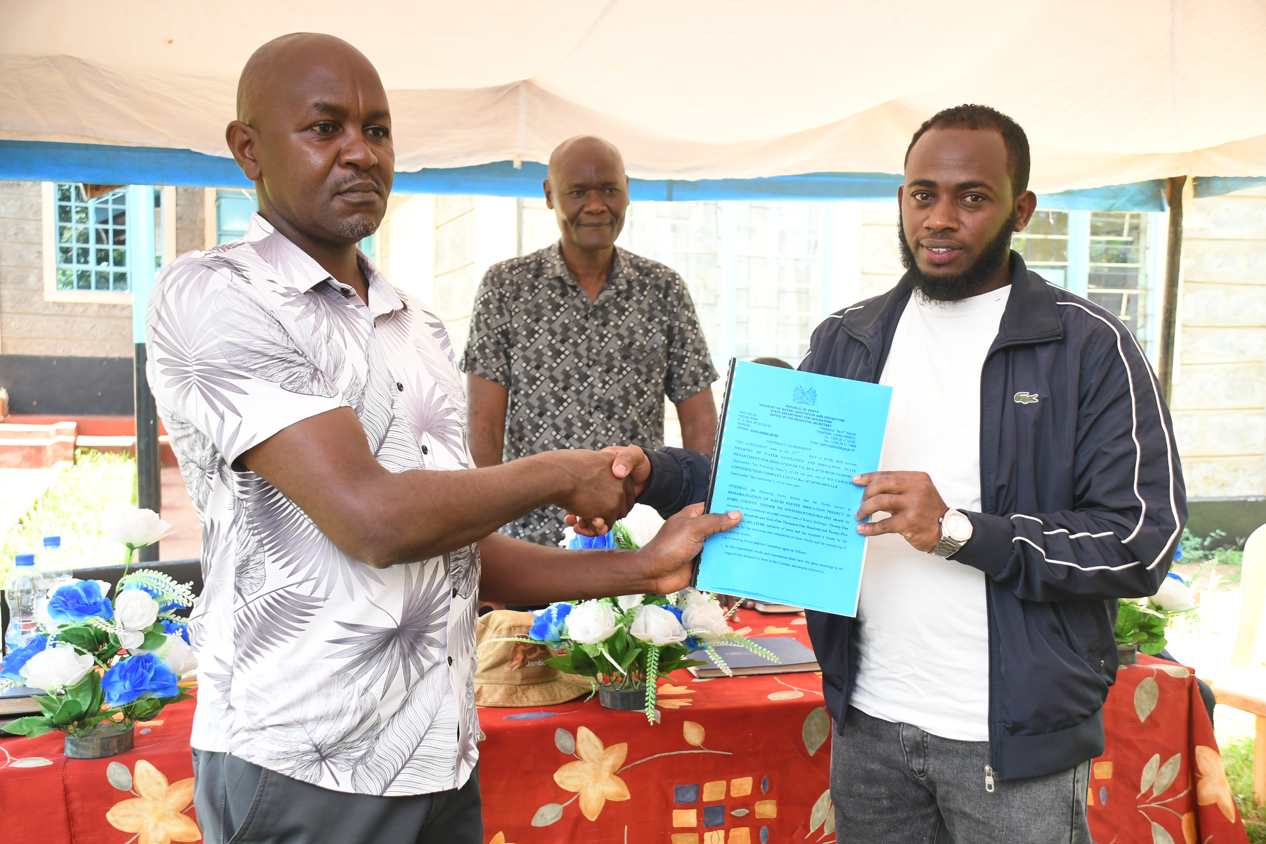 The Chairman Kiruki Kiende Farmers Cooperative Society, Bonface Kinyua Njiru (left), hands over the project contract documents for Kiruki Kiende irrigation project to Abubakar Mohamed from Gamachu Construction Limited (Contractor) as Director Irrigation Water Management,State Department for Irrigation, Daniel Odero looks on during the official site handover meeting at Kiruki Kiende Irrigation Water Users Association (IUWA) Offices in Gaturi South, Embu West Sub-County, Embu County.